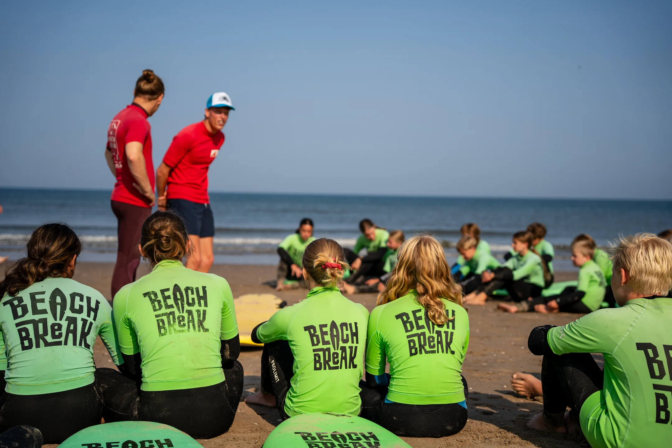 Een groep deelnemers in wetsuits krijgt uitleg van de coaches op het strand.