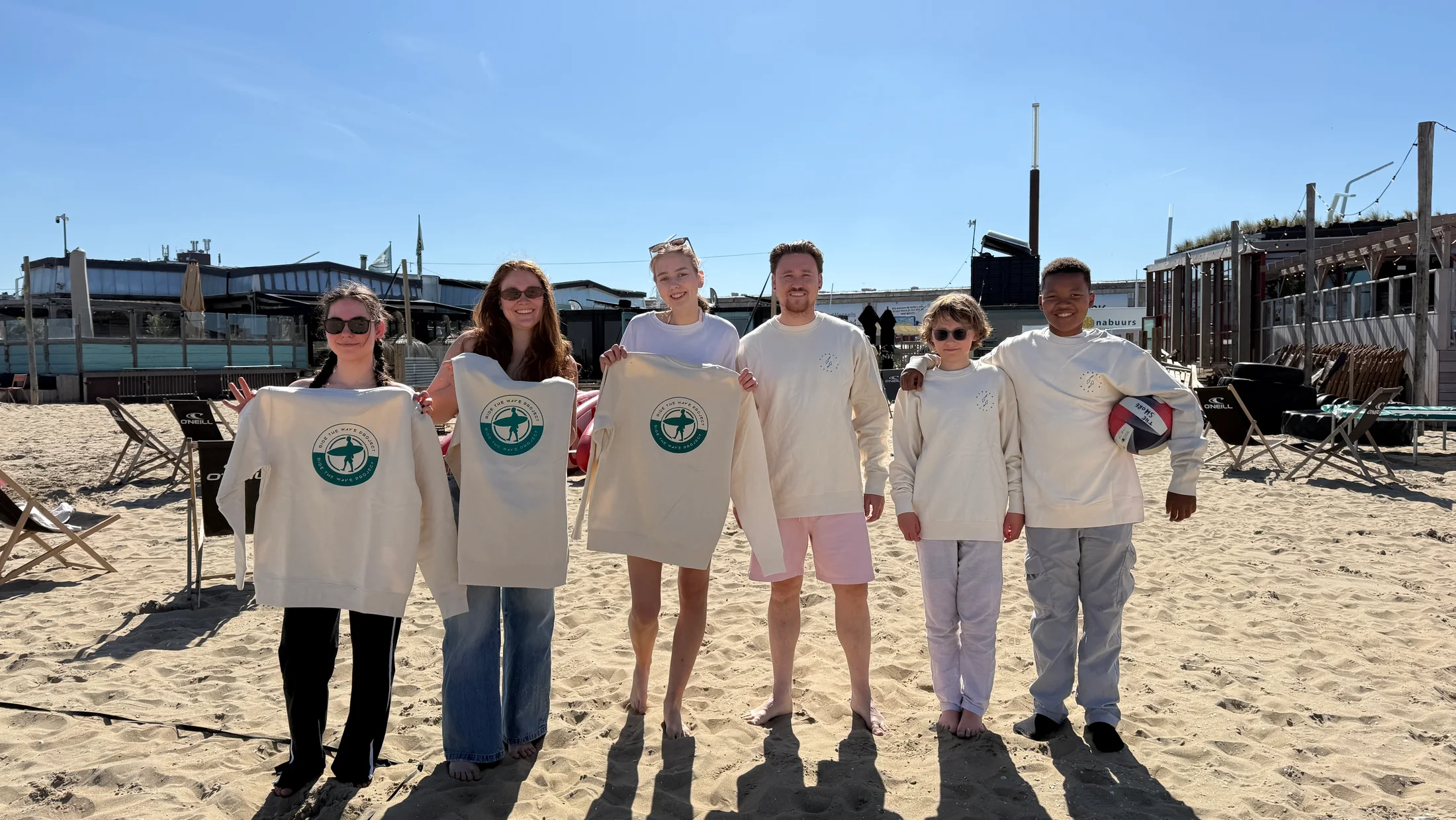 Een groep jongeren en begeleiders staat met Ride the Wave-kleding op het strand.