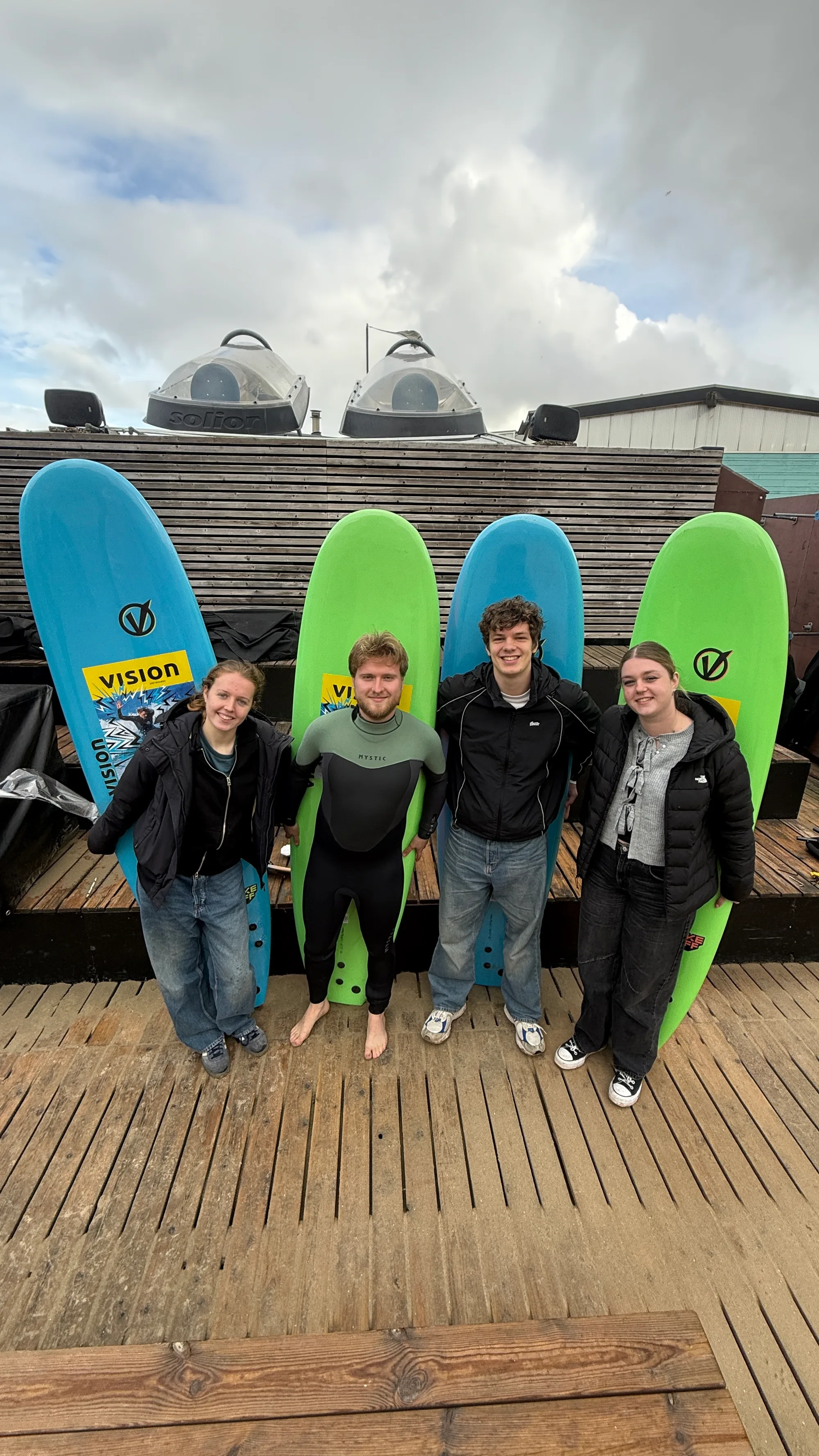 Vier jongeren staan met surfboards bij een surflocatie.