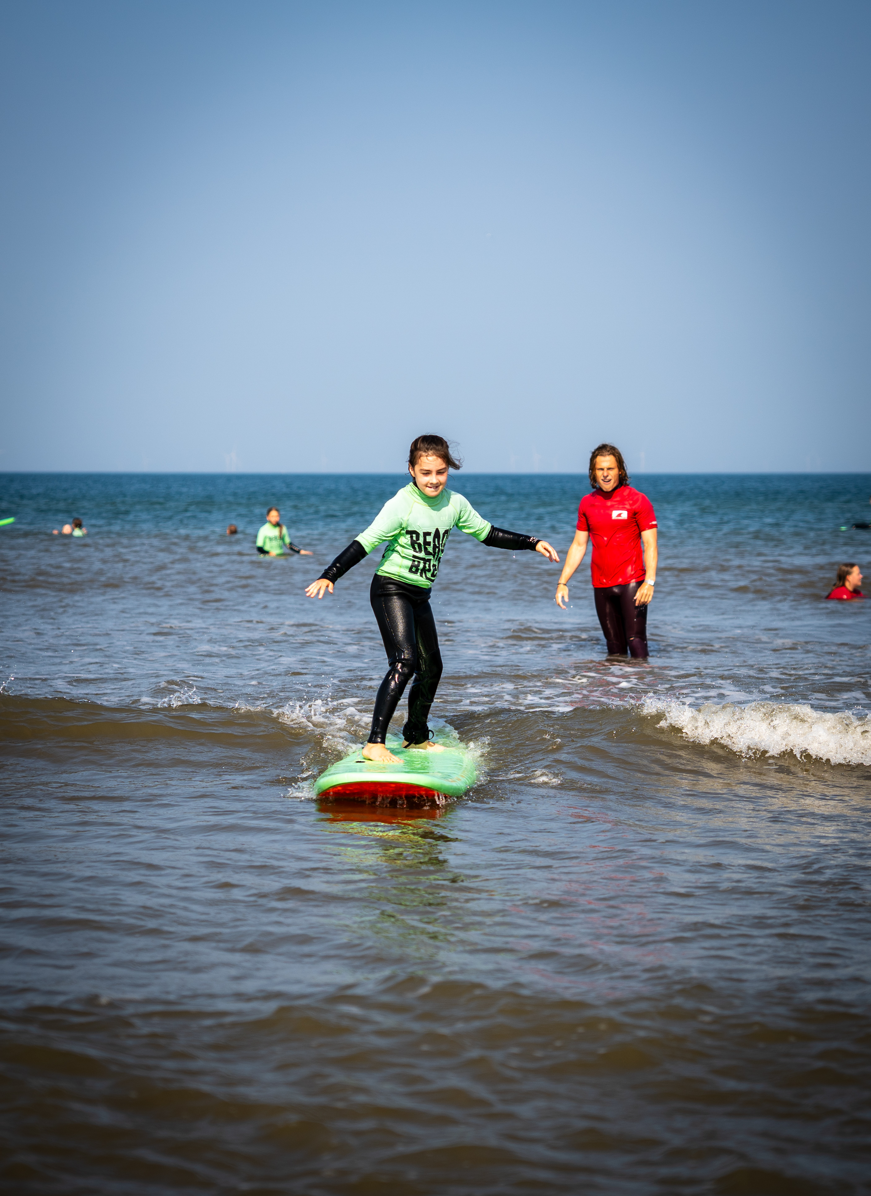 Een jonge deelnemer surft op een groene plank terwijl een coach meekijkt.