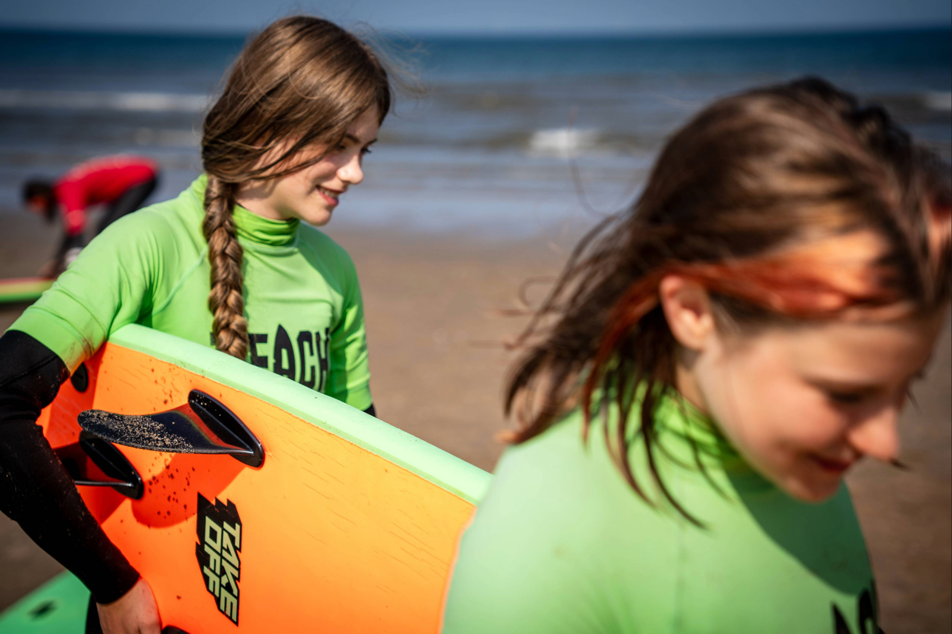 Twee jonge deelnemers lopen met een felgekleurd surfboard over het strand.